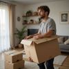 Man holding a cardboard box in a living room, looking thoughtful. Boxes are stacked on the floor.