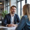 Man in a suit smiles and talks to a woman at a table in an office setting.