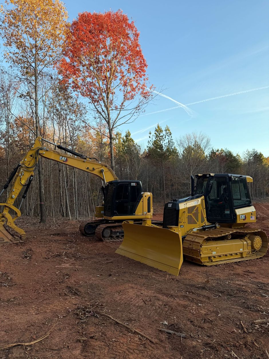 Yellow excavator and dozer on a dirt lot, clearing land for construction with trees in the background.