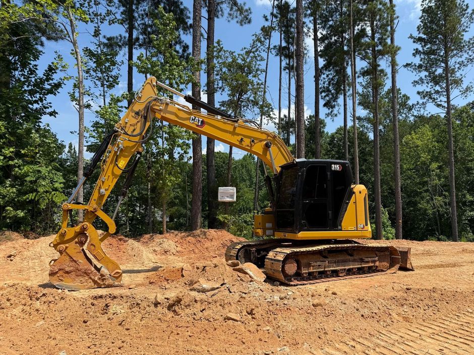Yellow excavator on a dirt lot, clearing land for construction with trees in the background.