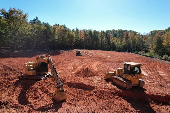 Construction site with yellow excavators on red dirt, trees in background, blue sky.