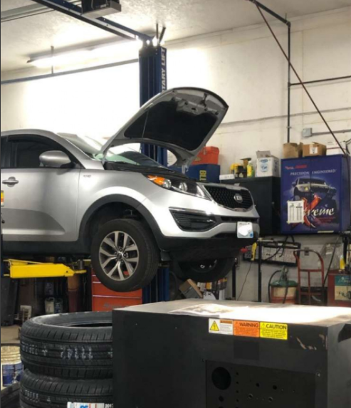 Silver Kia SUV on a lift with hood open in a repair shop. Tires stacked in foreground.