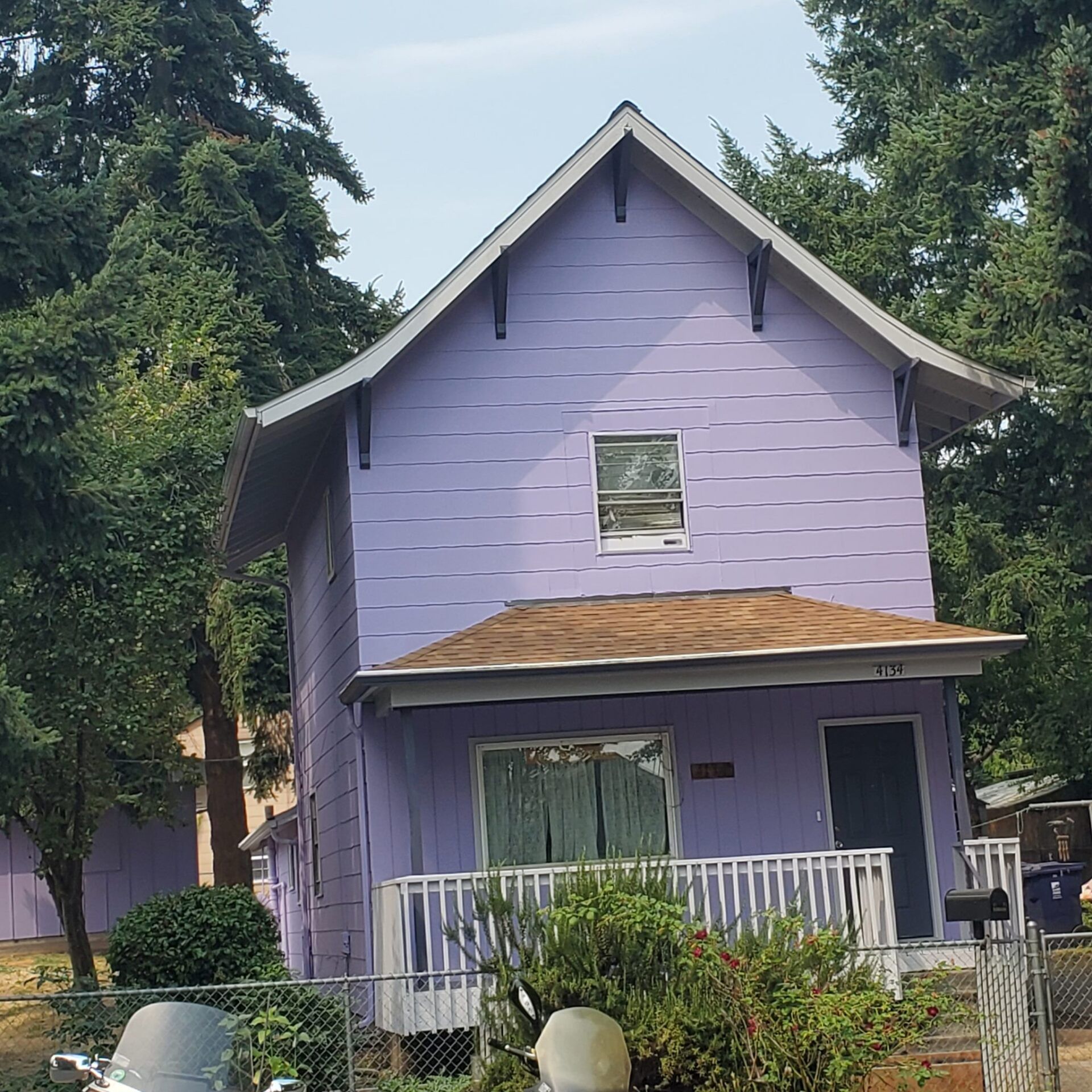 Two‑story house painted light purple with a small white porch and tan shingle roof, surrounded by tall evergreen trees.