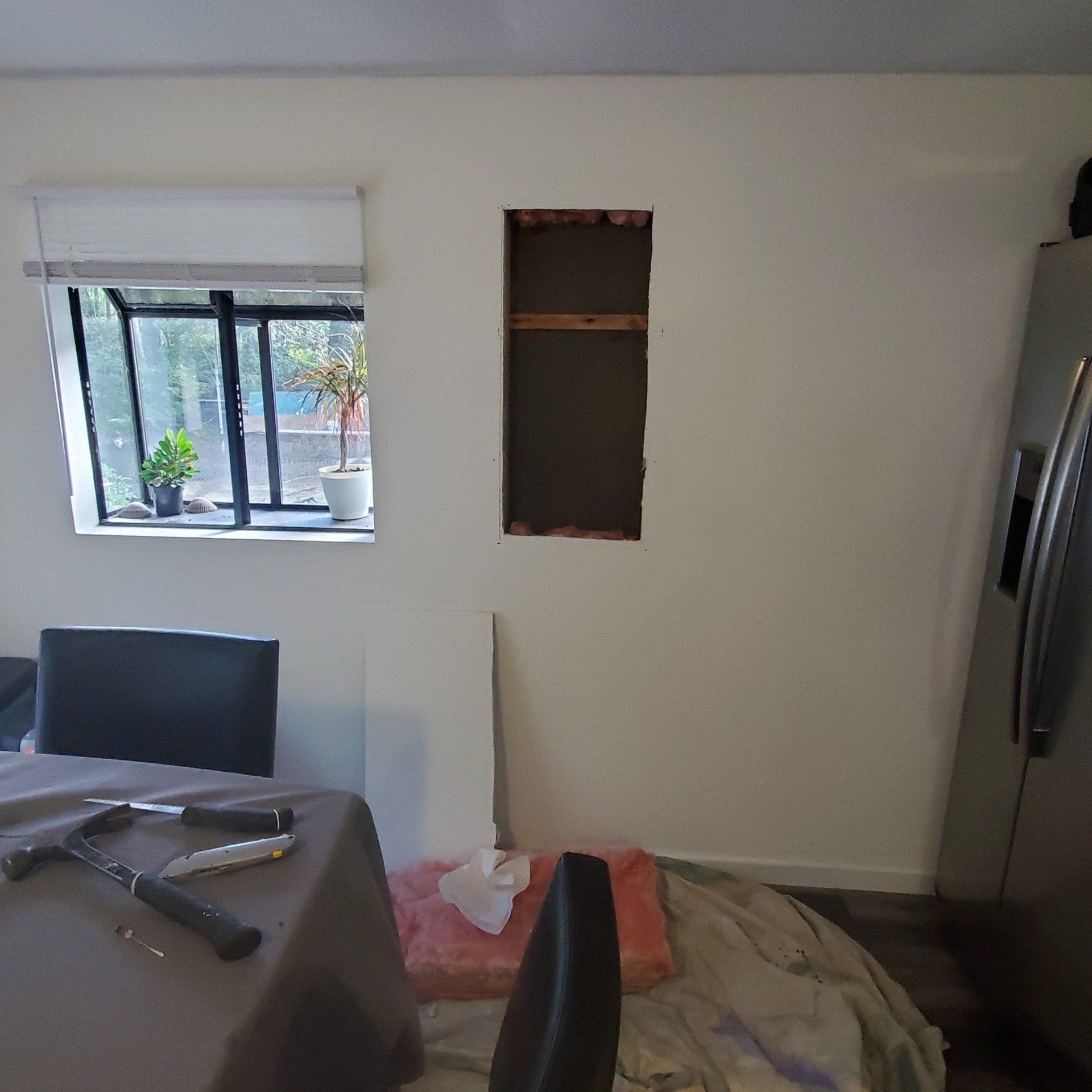 Wall with a rectangular hole cut between studs, tools and insulation on the floor, and a window with potted plants nearby.