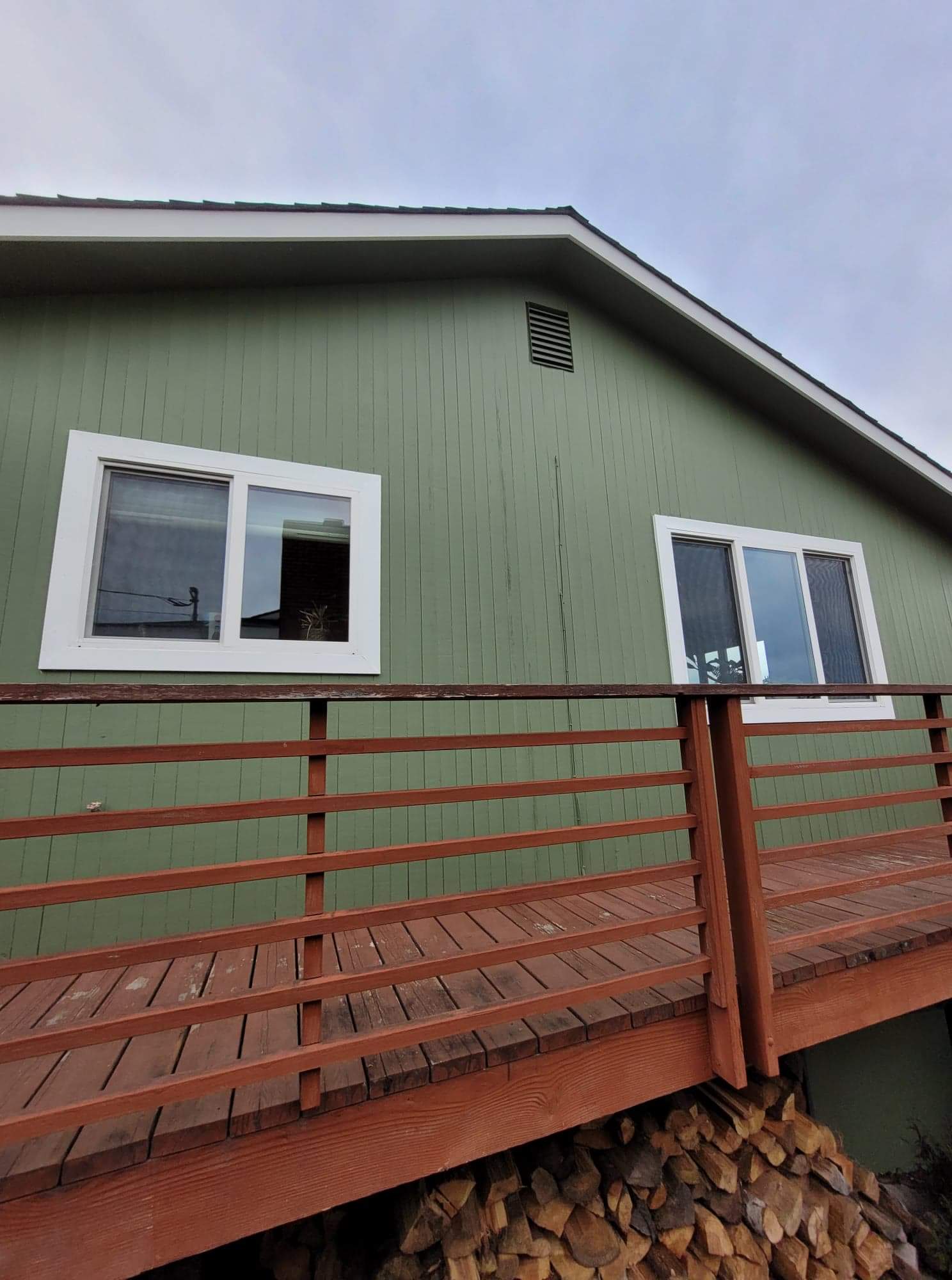 Side of a house with fresh green paint and clean white trim, next to a red‑brown deck with stacked firewood underneath.