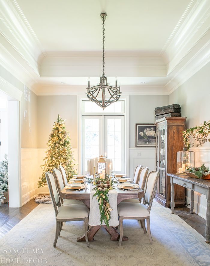 A bright white dining room with a set able, Christmas tree, chandelier, and a decorative cabinet.