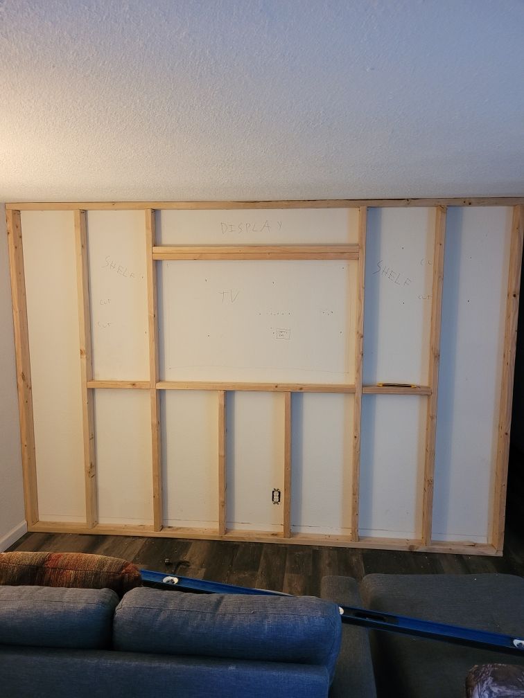 Unpainted framed wall with built‑in sections for a TV, display shelf, and shelving, under construction in a living room.