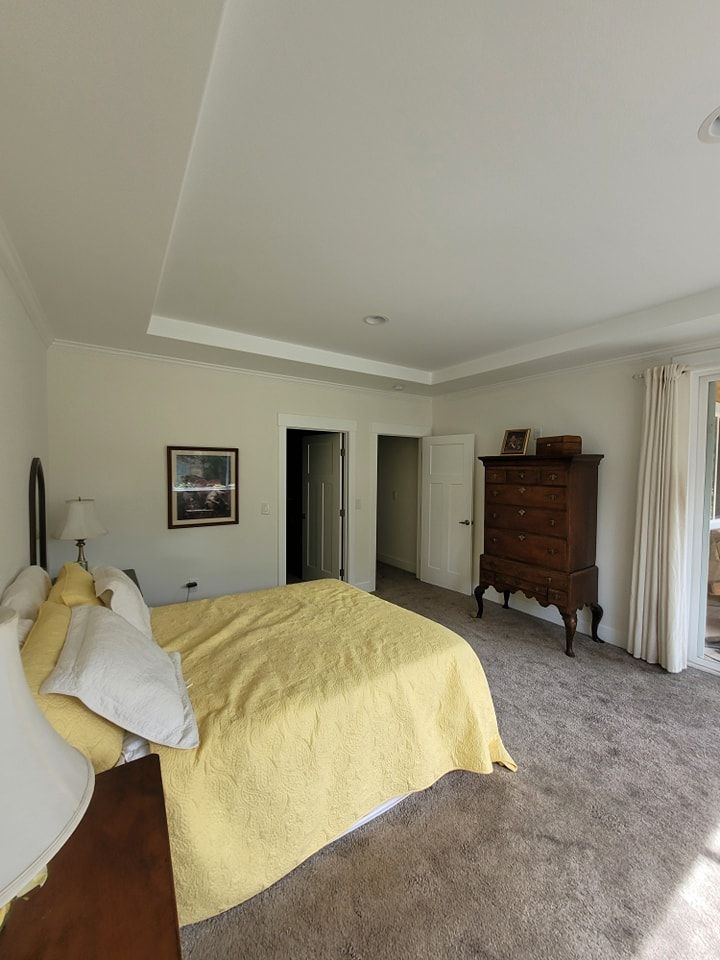 Bedroom with beige walls, carpeted floor, a bed with yellow bedding, and a tall wooden dresser near the window.