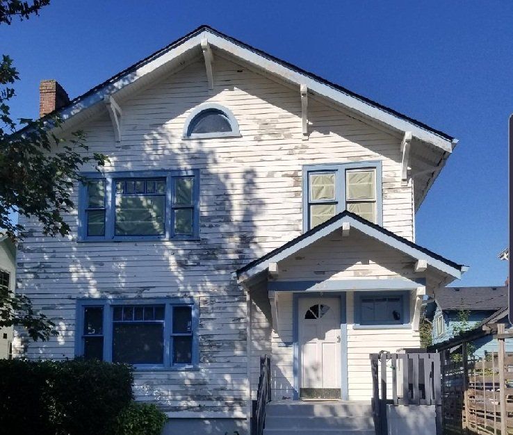 A weathered two‑story white house with an arched window, blue window frames,and peeling paint.
