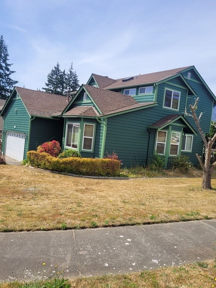 Two‑story house with dark green siding, light green trim, and a brown shingle roof on a dry lawn.