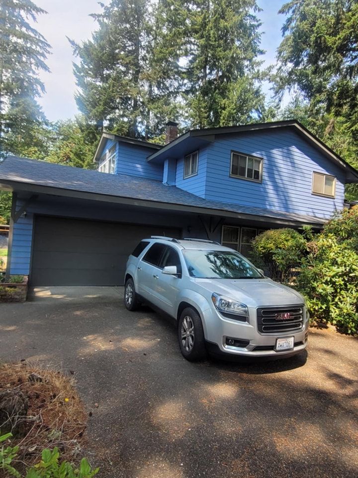 Blue exterior siding with dark blue trim and garage door, with a car in the driveway and surrounding pine trees