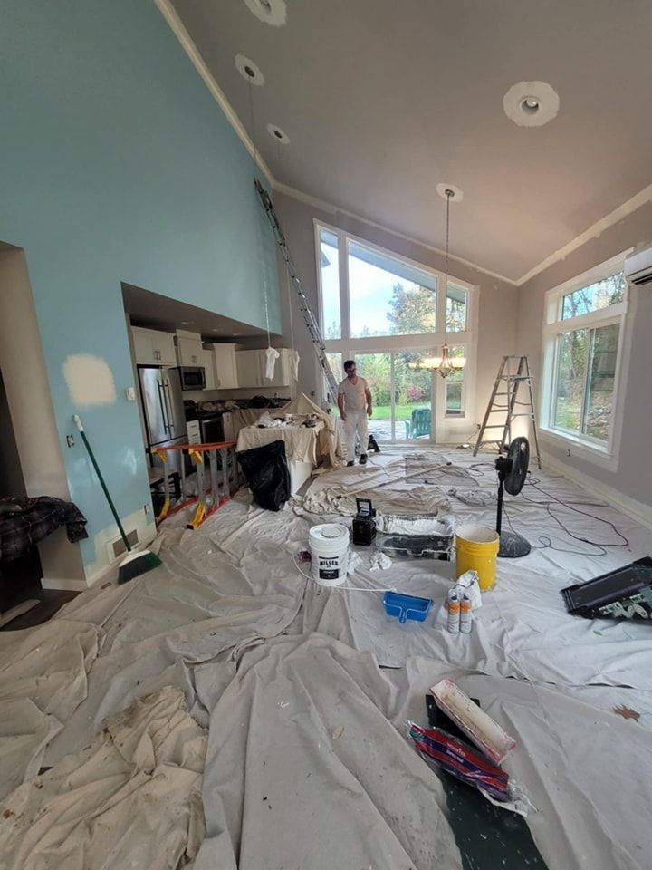 Interior room being painted, blue wall, painter on ladder, white paint, drop cloths, supplies on floor.