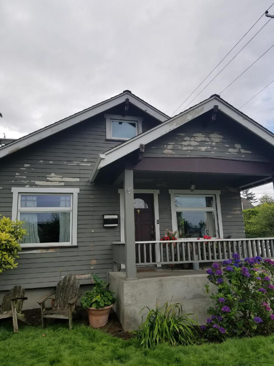 Gray house with white‑framed windows, a porch, and peeling siding paint.