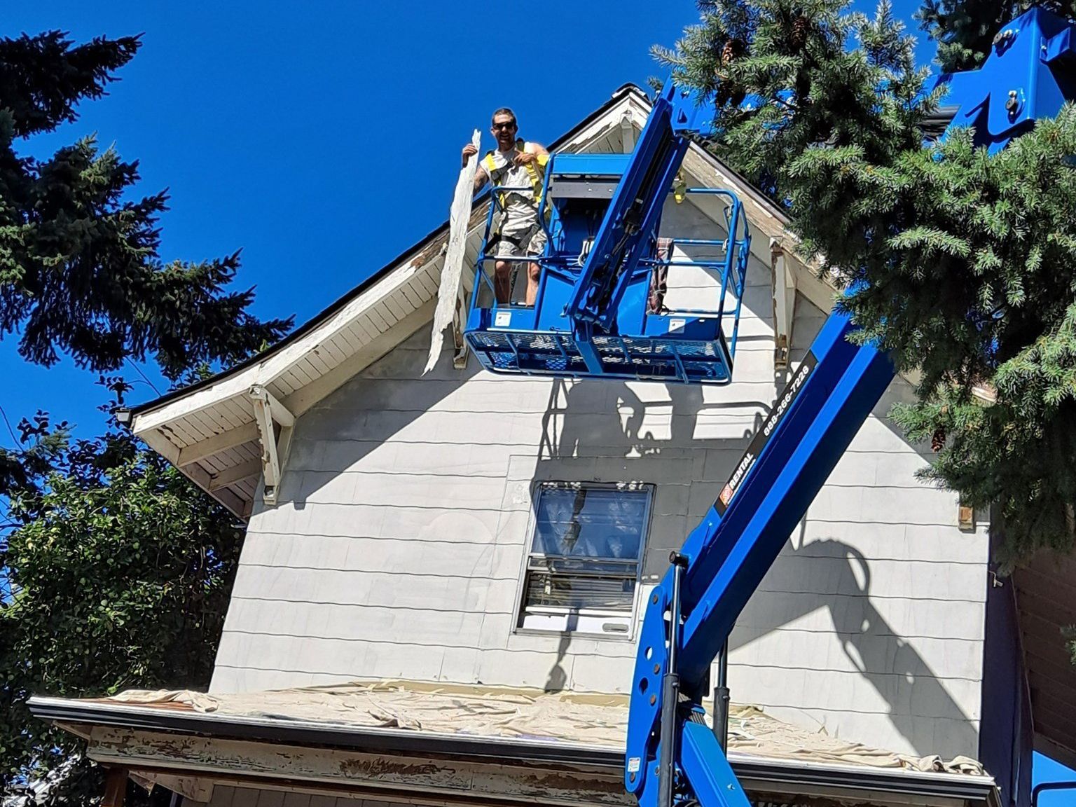 A Paint-Right Painting worker on a lift removing old siding from a tall two-story house with a wood porch and white exterior.