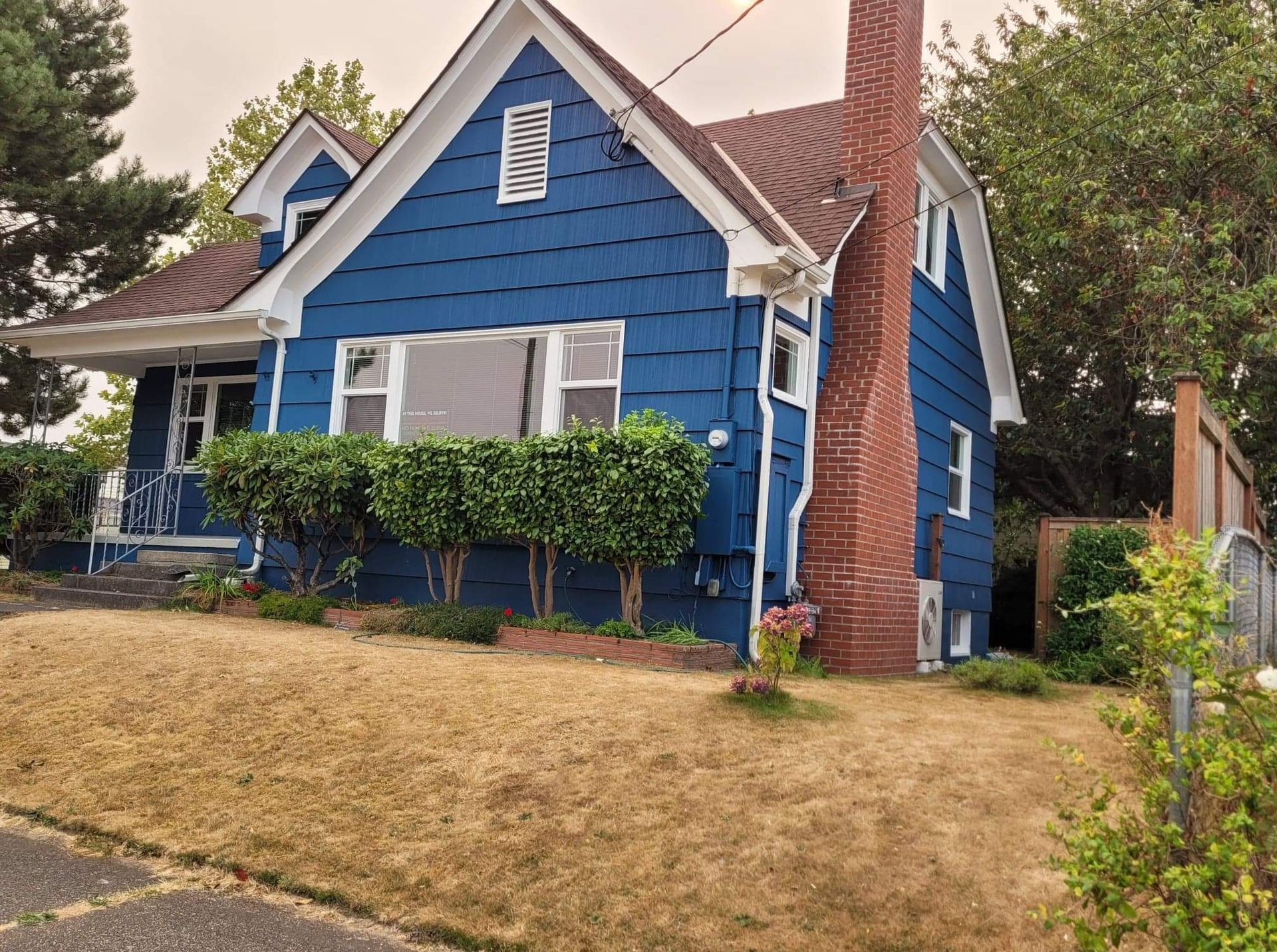 House with deep blue painted siding, white trim, and a red brick chimney, set above a sloped lawn and trimmed hedges.