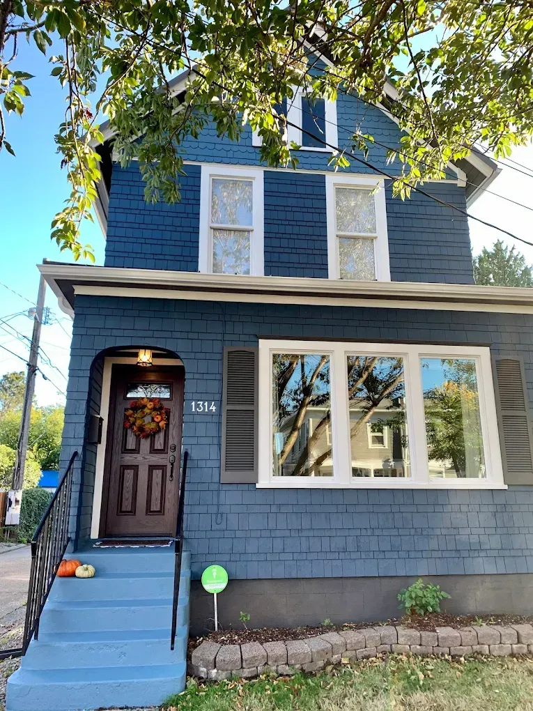 Blue house with brown door and white-framed windows, porch with pumpkins, under a tree on a sunny day.