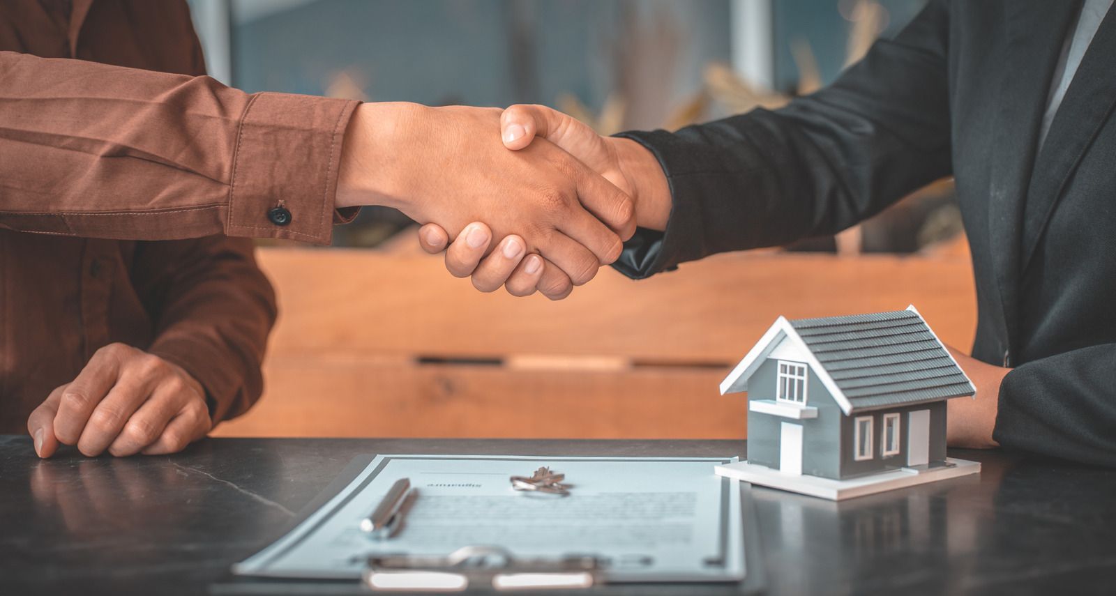 Two people shaking hands over a table with a small house model and keys, suggesting a real estate deal