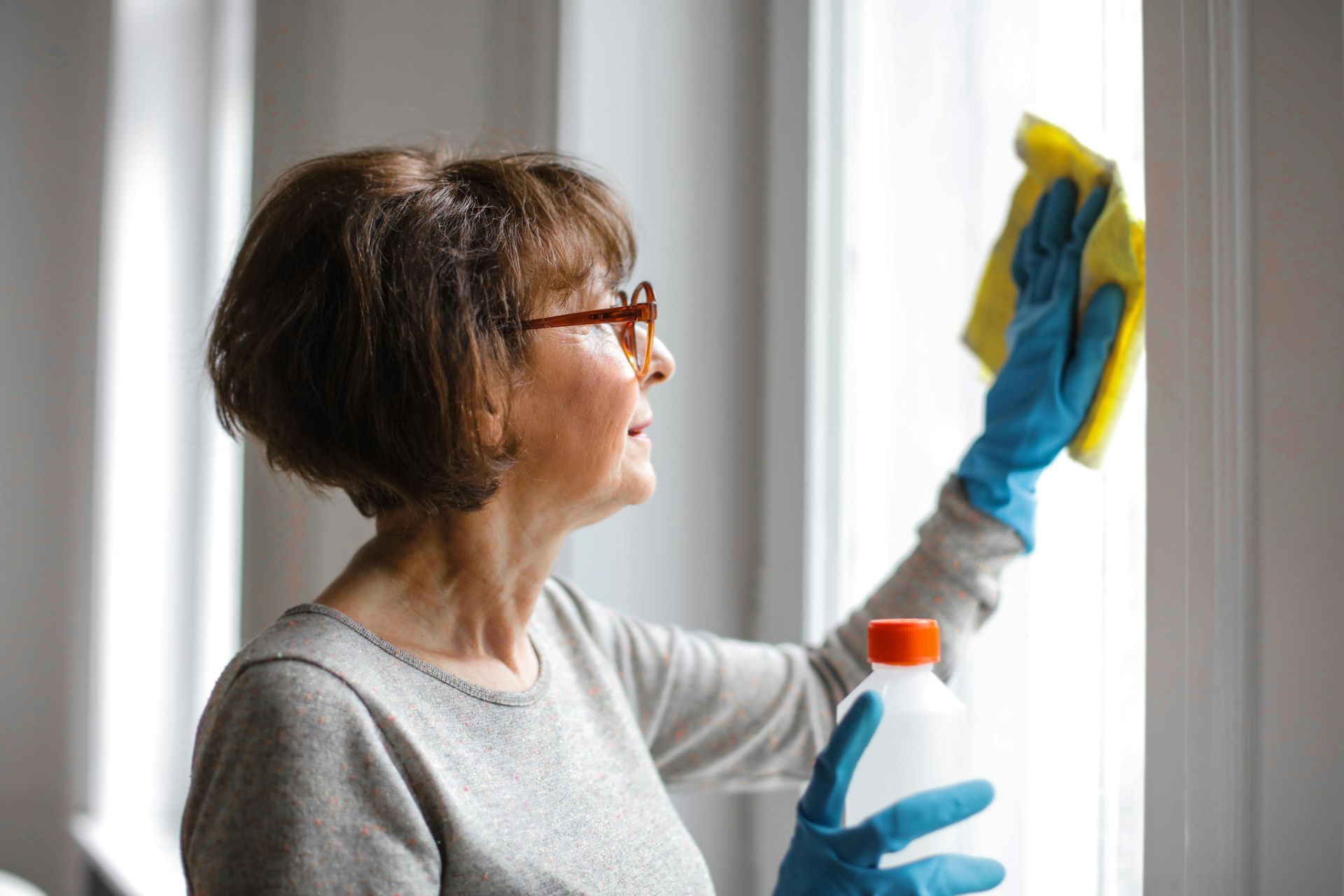 Person cleaning a window with a spray bottle and cloth, wearing blue gloves and glasses