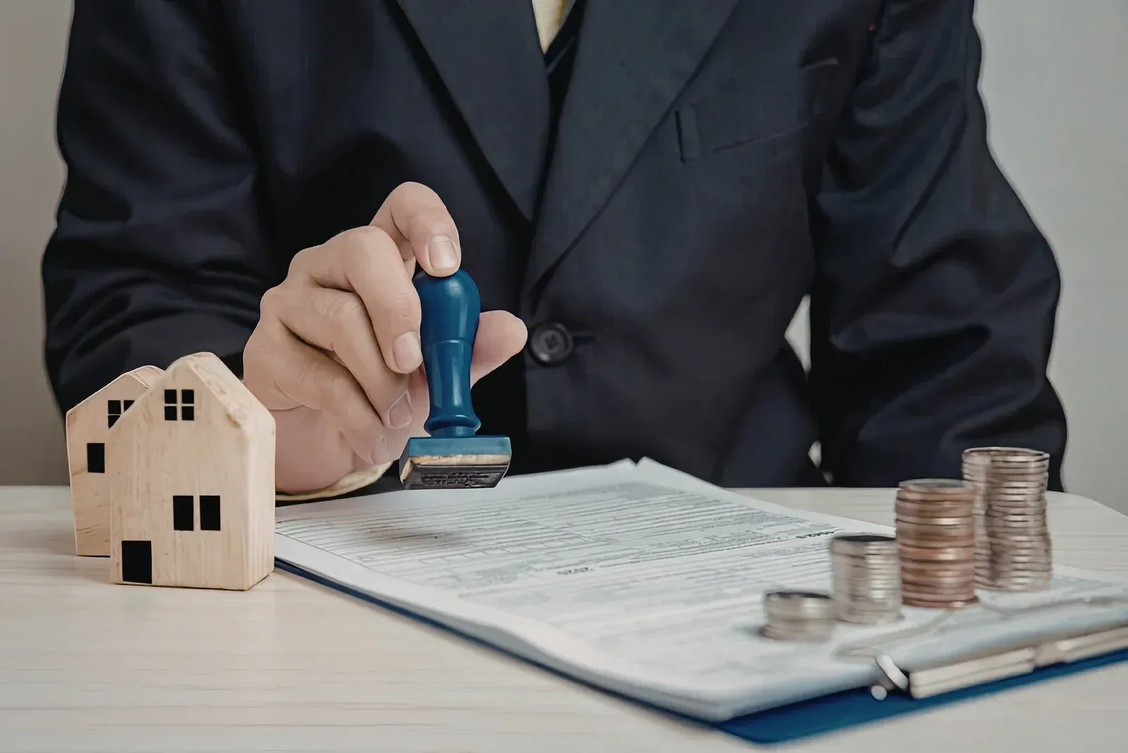 Person stamping paperwork beside a small house model and stacked coins, suggesting property appraisal or mortgage processing