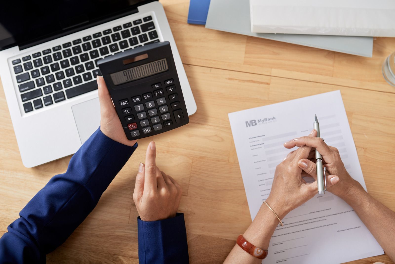 Hands using a calculator and writing on a form at a desk with a laptop