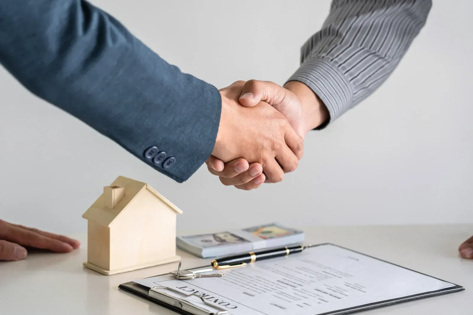 Two people shaking hands over a house model, paperwork, and a pen on a desk