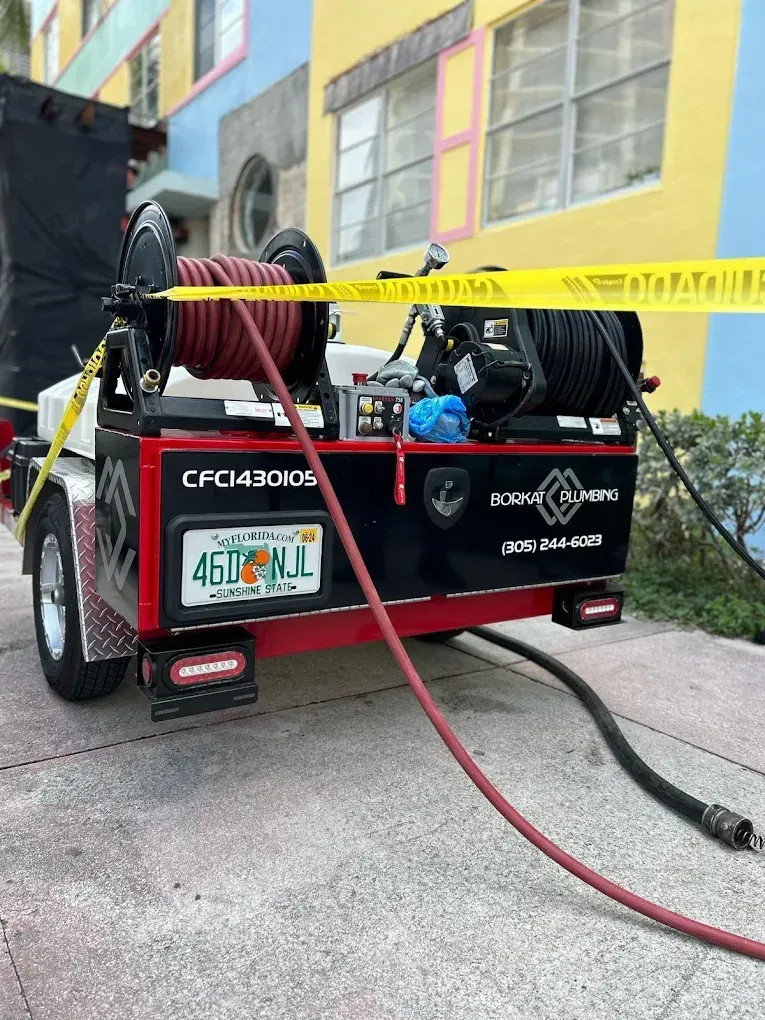 A red and black trailer with coiled hoses sits in front of a colorful building, marked by caution tape, likely used for cleaning or repairs.
