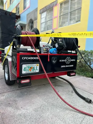 A red and black trailer with coiled hoses sits in front of a colorful building, marked by caution tape, likely used for cleaning or repairs.