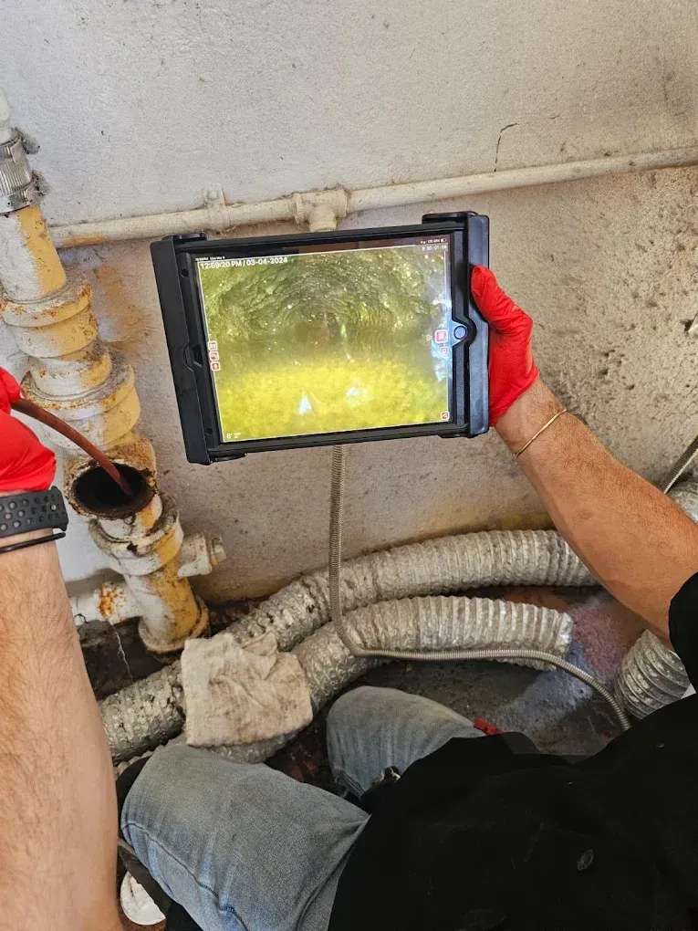 A person uses a camera to inspect a pipe; the screen shows the inside. Red gloves and denim jeans are visible.