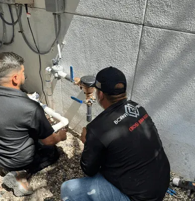 Two men in black shirts inspect outdoor plumbing near a light-colored wall. One kneels, the other sits.