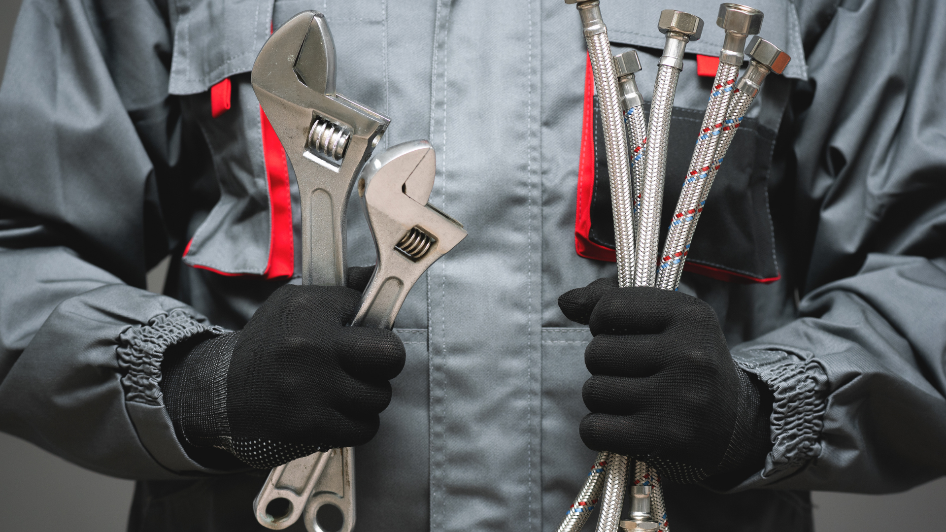 Person in workwear holding two adjustable wrenches in one hand and a bundle of flexible plumbing hoses in the other.