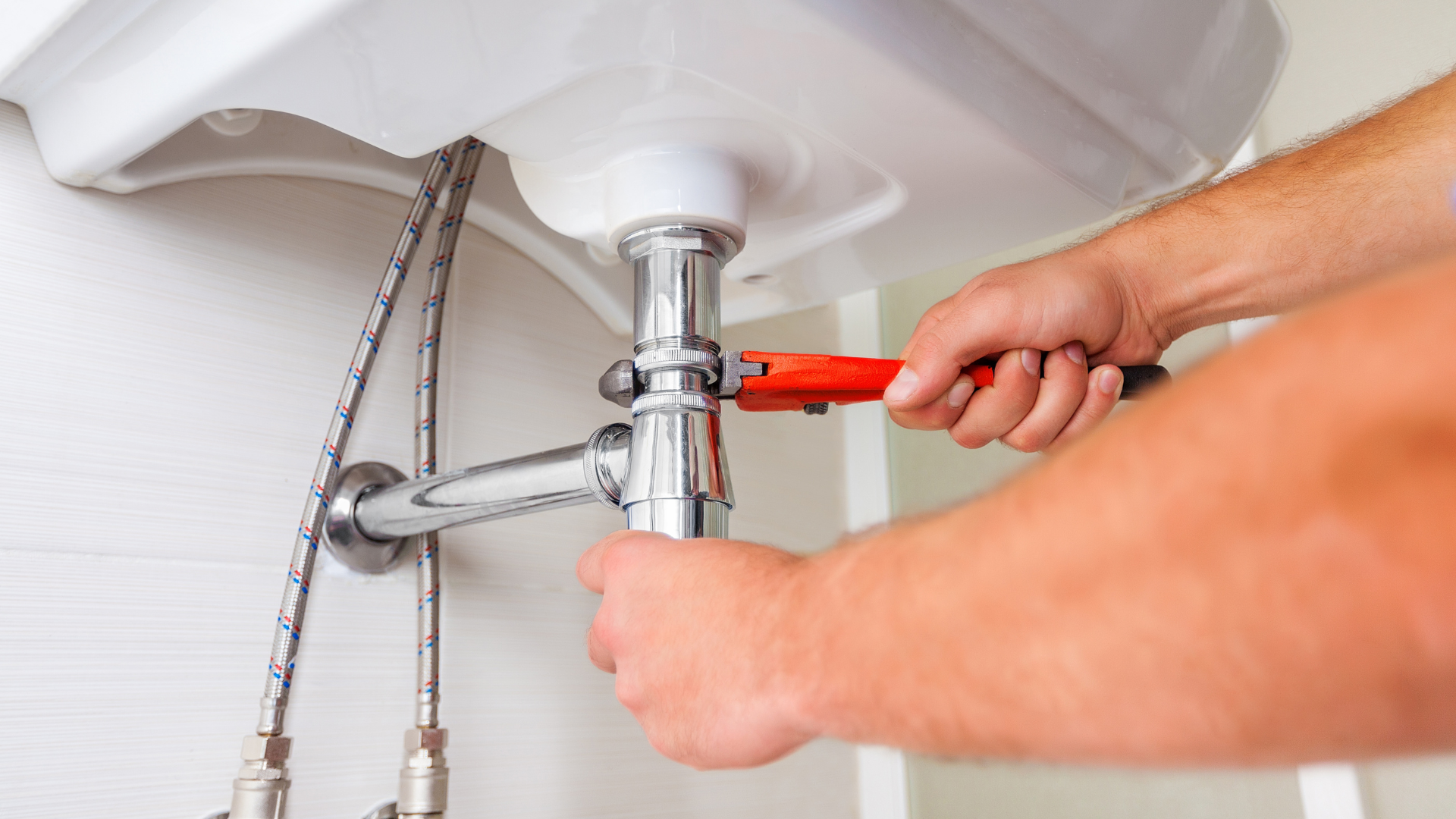 Plumber using a wrench under a white sink. He is tightening a pipe fitting, a chrome-colored drainpipe.