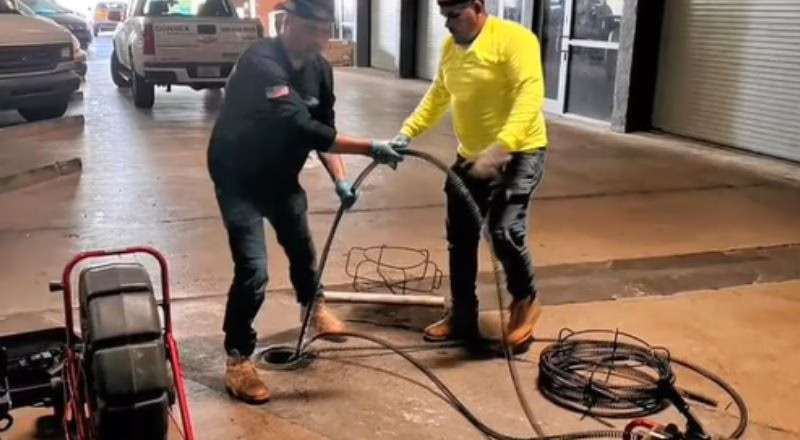 Two men in work clothes unclogging a drain in a garage. One holds a cable, the other assists. A truck is parked in the background.