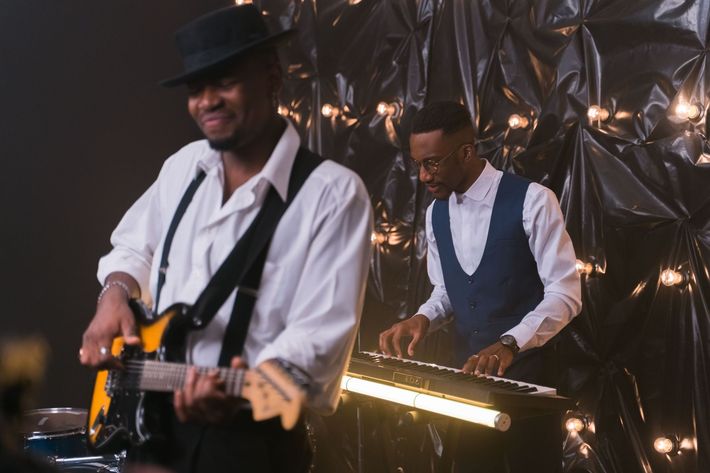 Two musicians performing on stage, one playing guitar and one using a keyboard, with warm lighting.