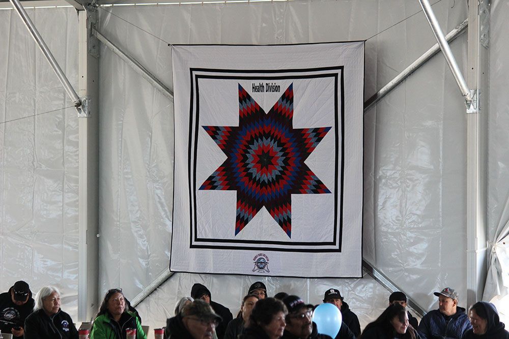 A group of people are sitting under a tent with a quilt on the wall.