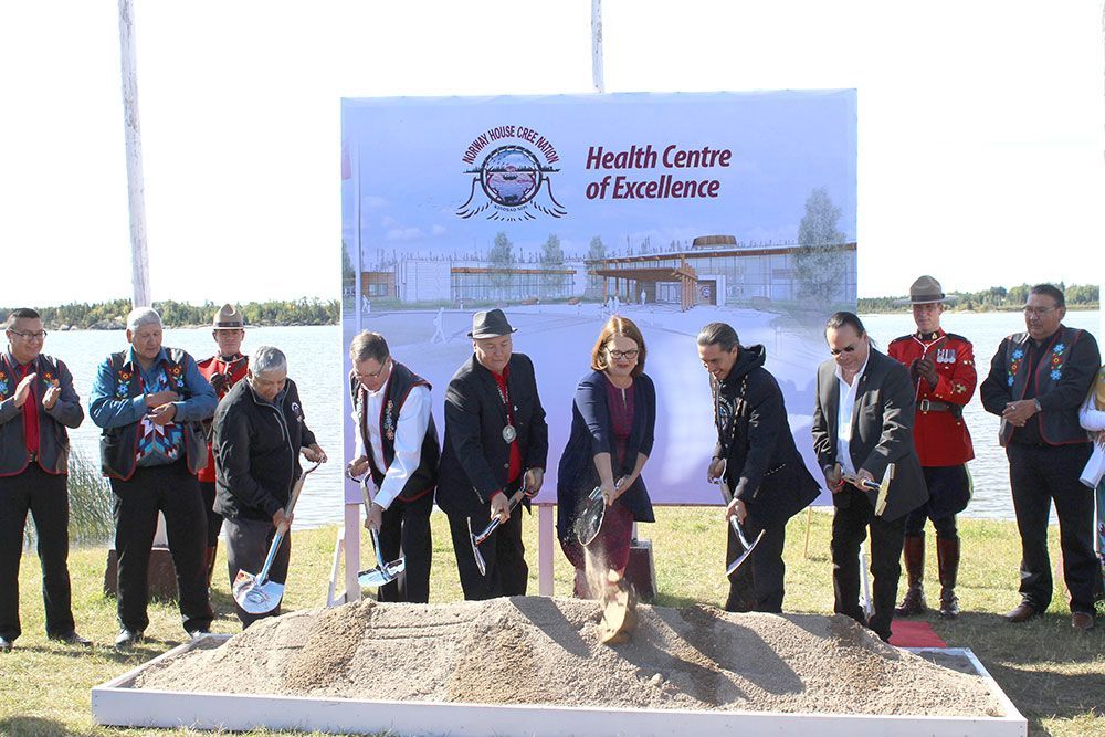 A group of people are shoveling dirt in front of a sign that says health centre of excellence
