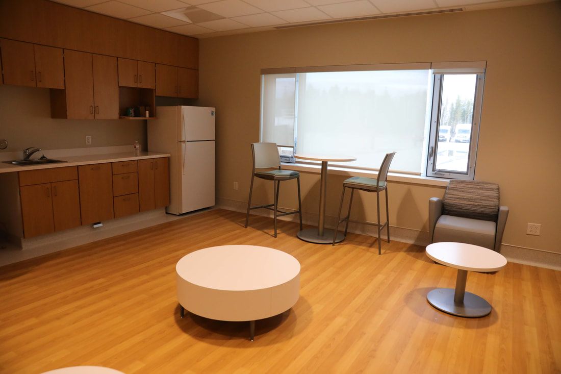 A living room with a refrigerator , chairs , tables and a window.