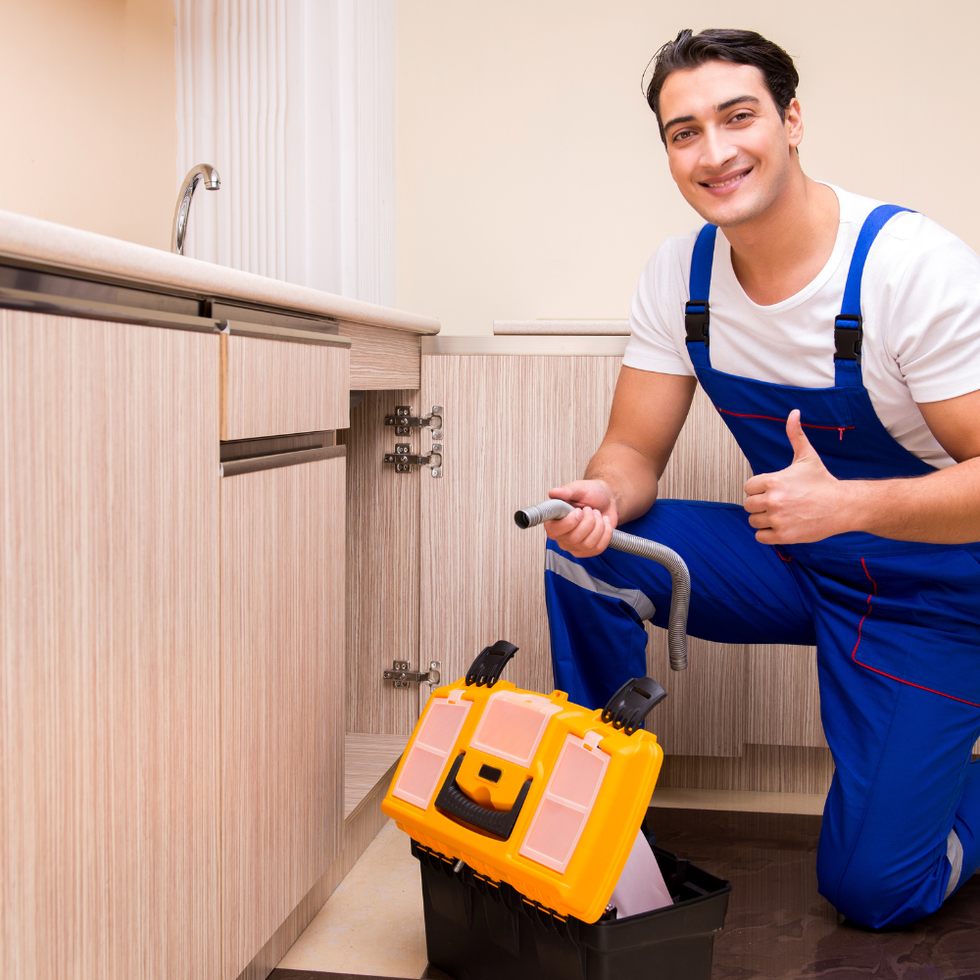 Plumber kneeling, smiling, and giving thumbs-up next to kitchen cabinet with tools.
