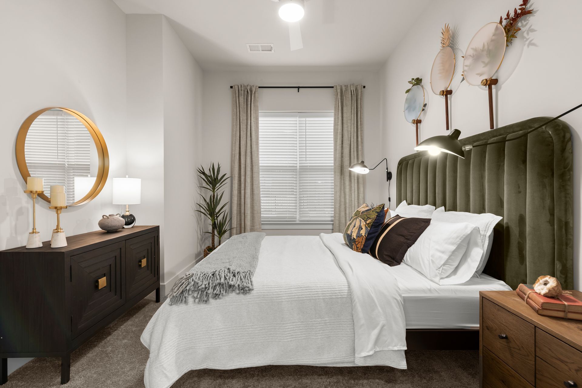 Bedroom with a velvet headboard, dresser, round mirror, window with blinds, and decorative wall art.
