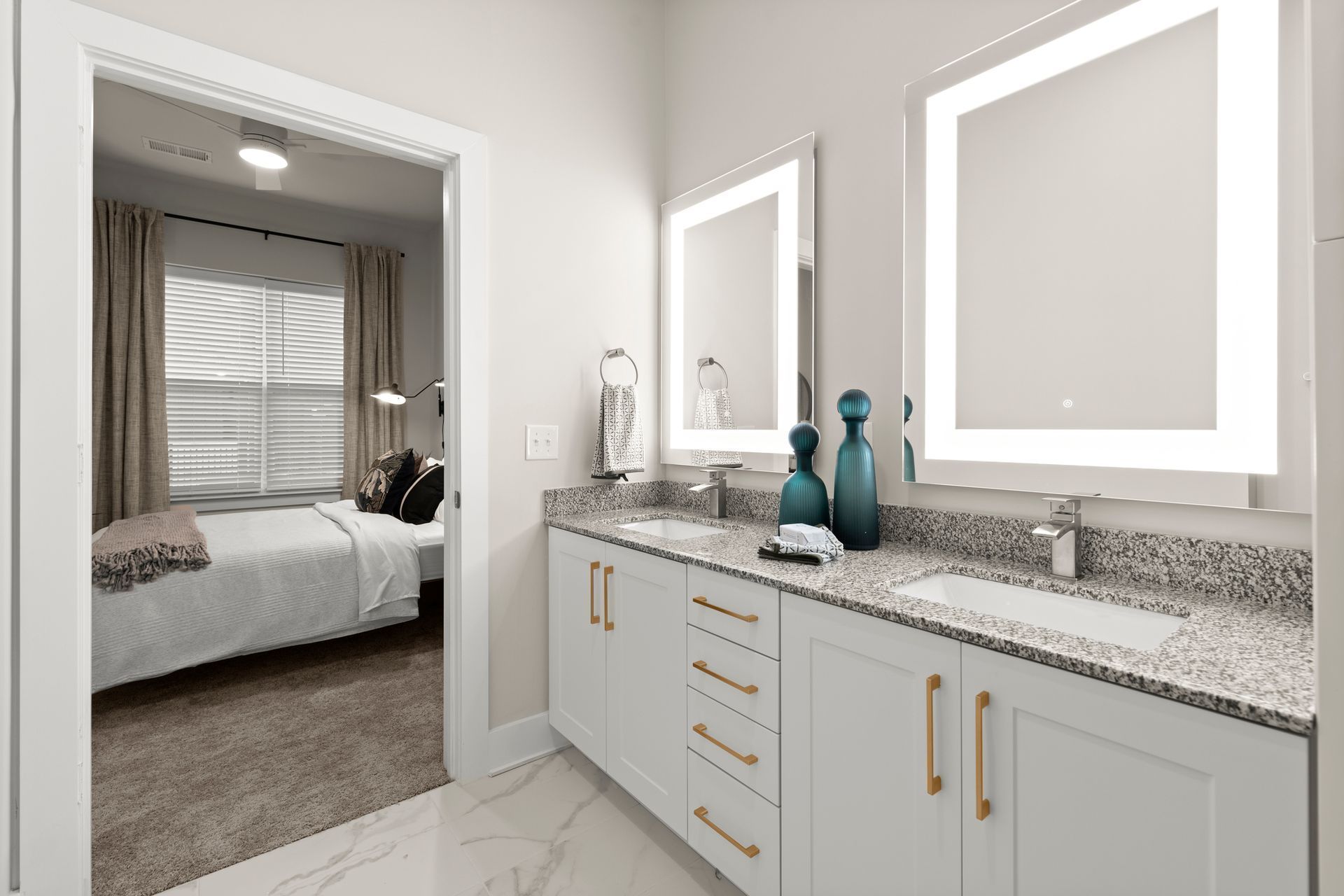 Bathroom with double vanity, lit mirrors, and door to bedroom. Light gray walls, white cabinets, and granite countertop.