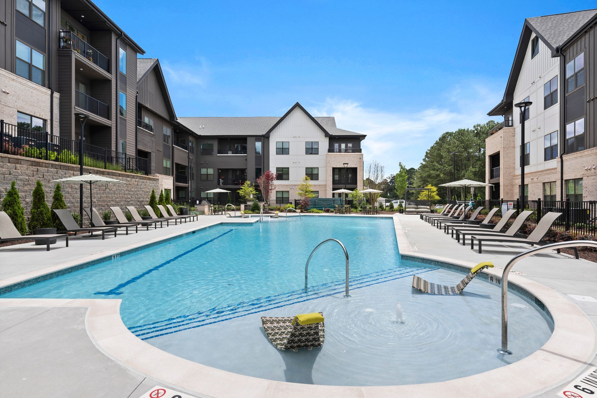 Swimming pool surrounded by lounge chairs, in front of modern apartments with a sunny, blue sky.