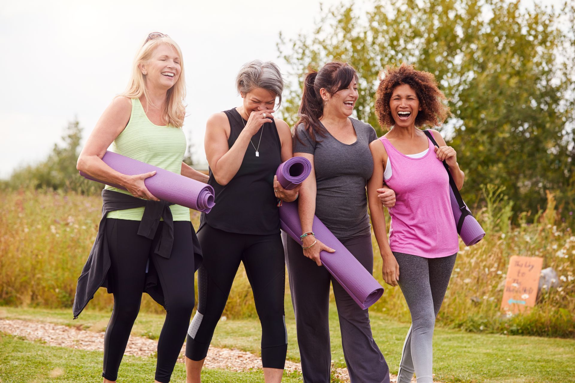 Four women in workout clothes laugh, holding yoga mats, walking outdoors in a grassy area.