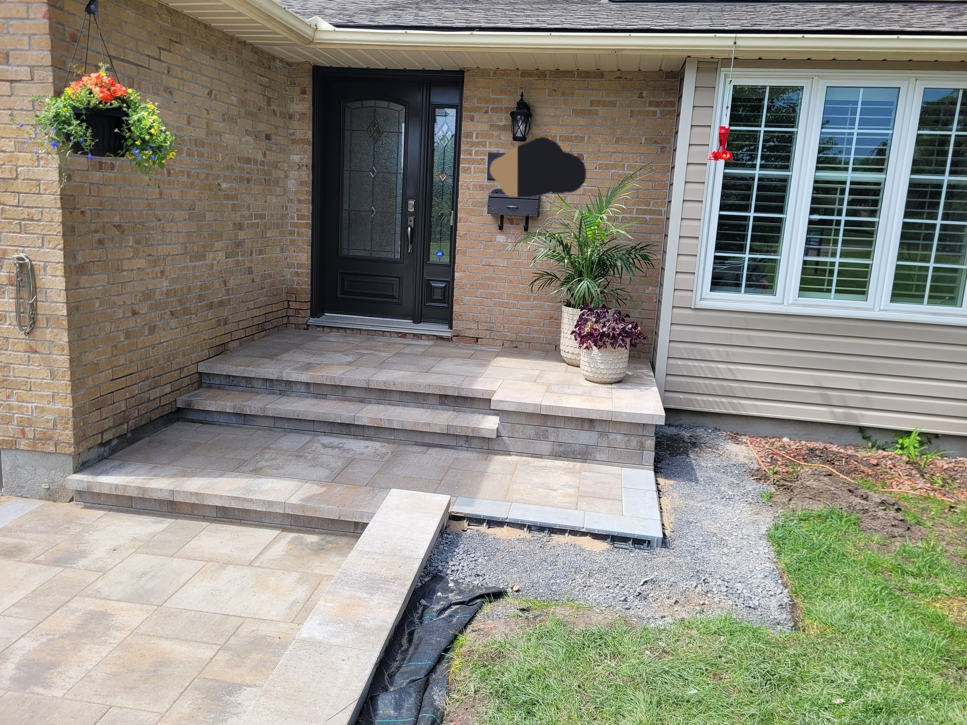 Brick house entrance with stone steps and walkway, black door, hanging flower basket, and a window.