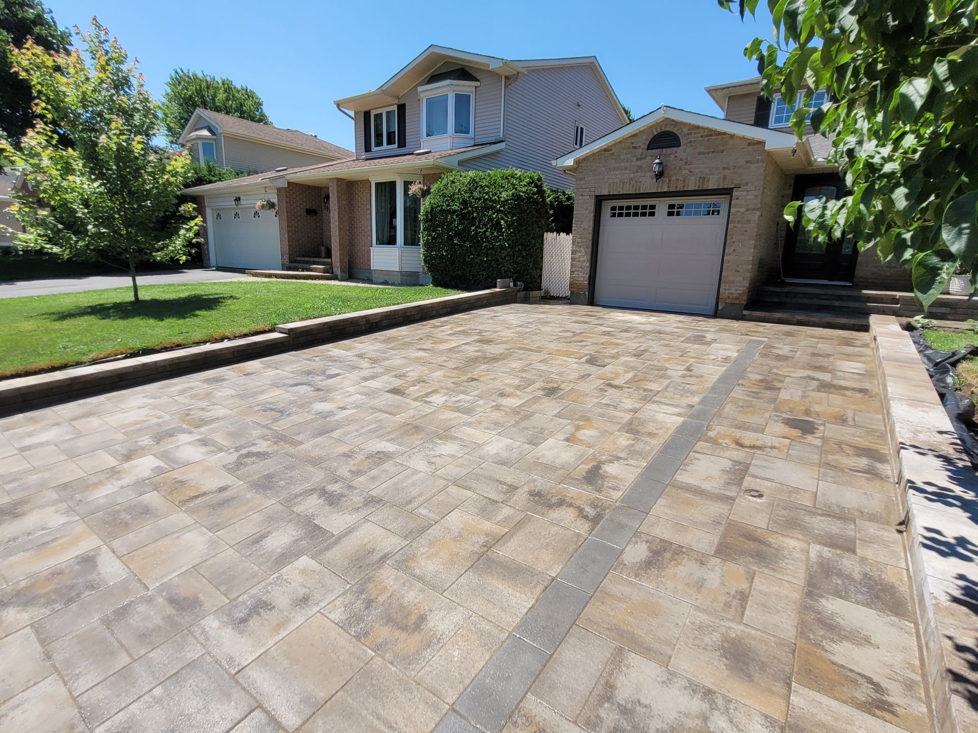 Driveway paved with tan and gray stones leading to a two-story house with garage on a sunny day.