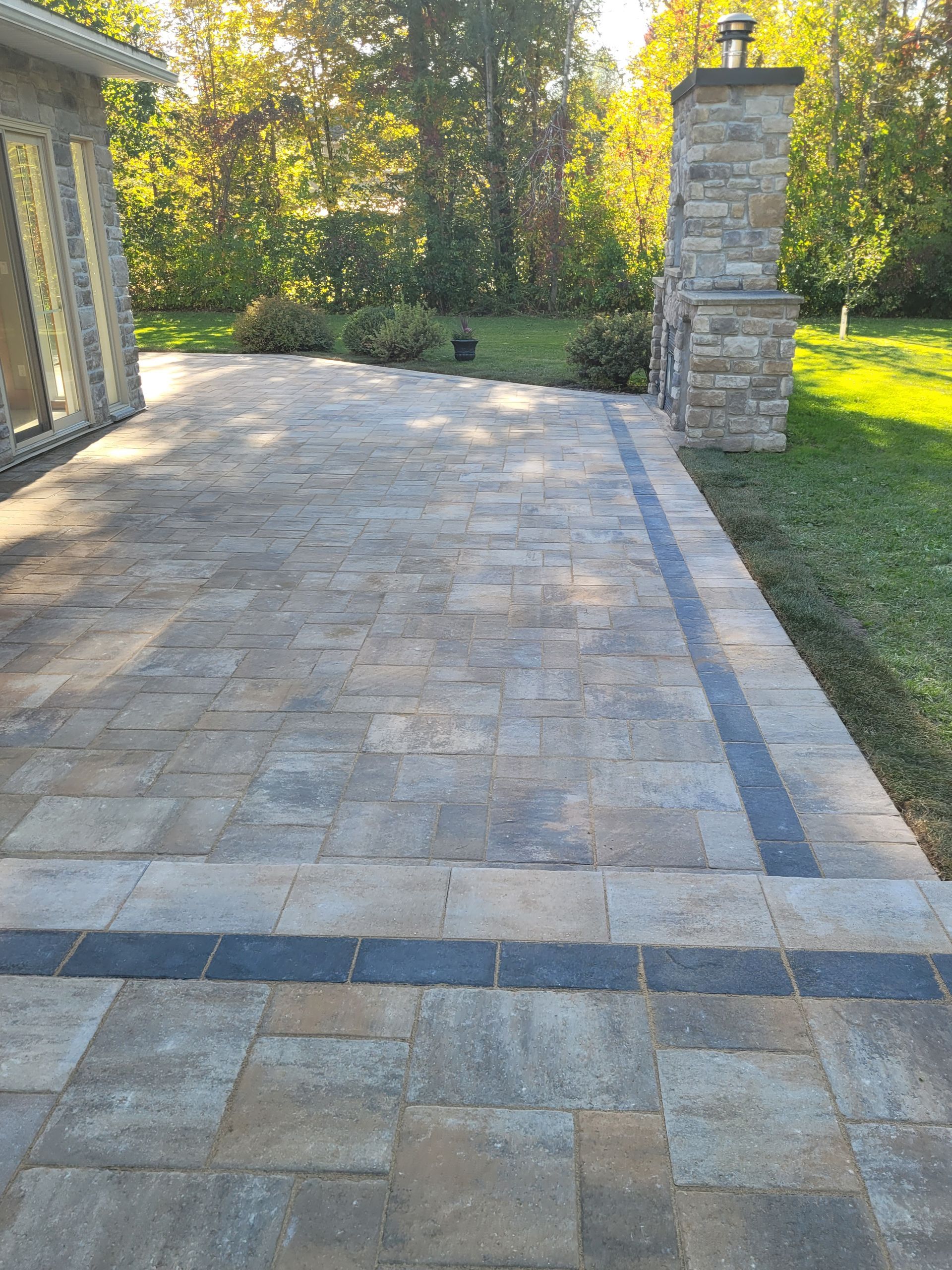 Stone patio with dark border, leading to an outdoor fireplace, green grass, and trees.