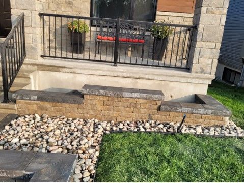 Stone steps leading to a house with a black railing, planters, and a bench; gravel and grass.