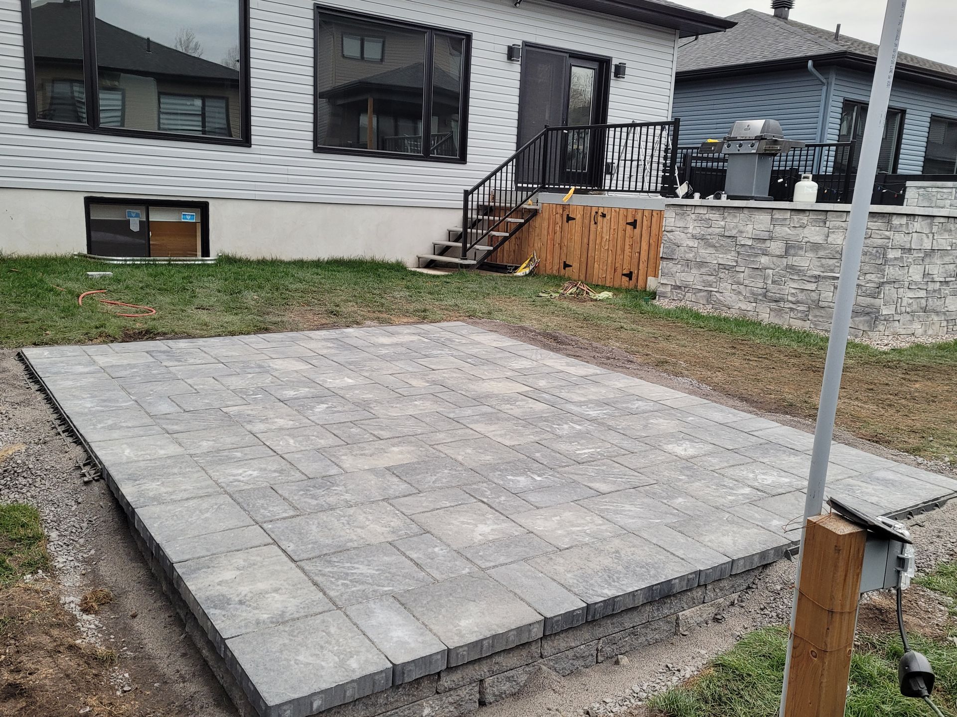 Patio pavers installed on a backyard lawn near a house with a raised deck and stairs.