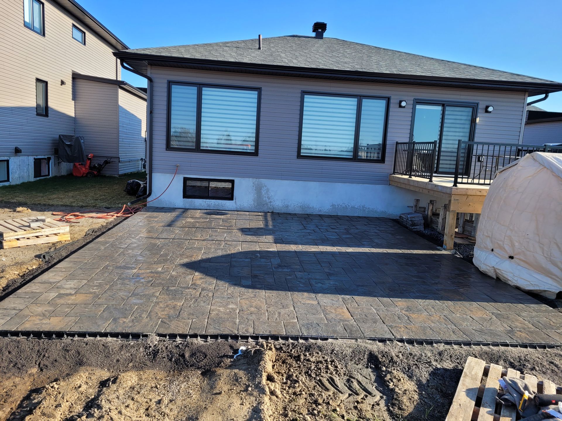 Backyard patio construction: gray pavers, house with windows, deck, blue sky.