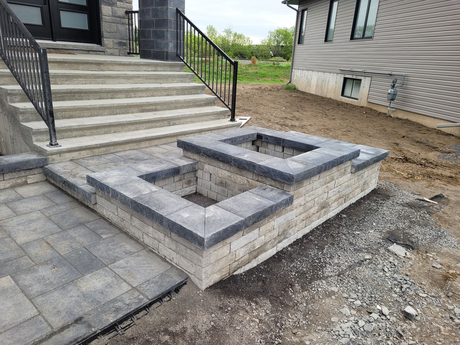 Stone steps and retaining wall lead to a house entrance with a black metal railing, outdoors.