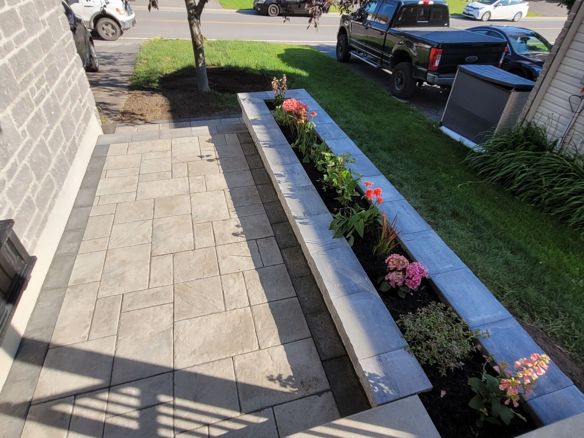 Patio en pierre grise avec parterre de fleurs et marches menant à une maison, entouré de verdure et d'un camion.
