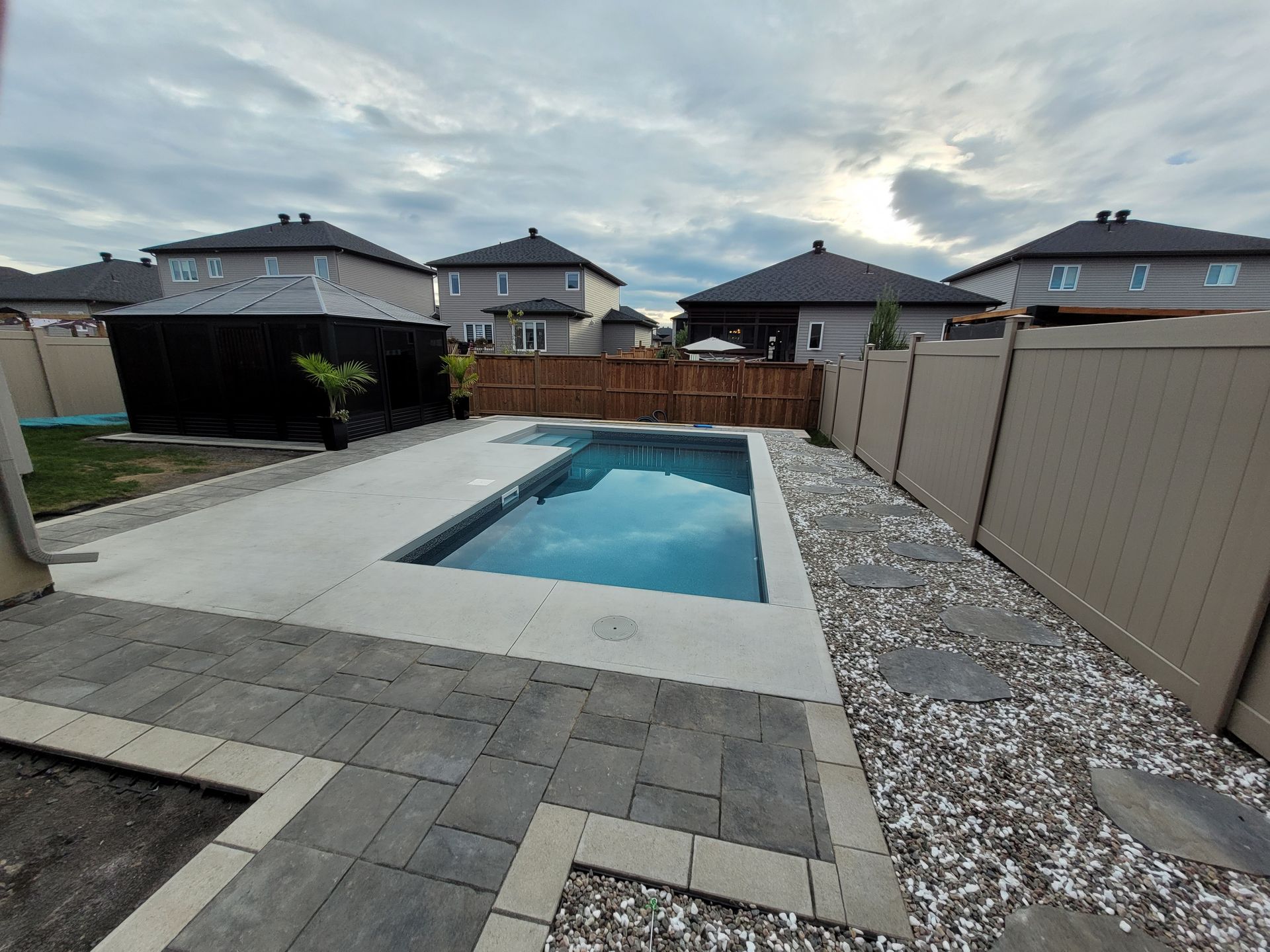 Backyard with a rectangular pool, patio, and fences. Houses in the background under a cloudy sky.